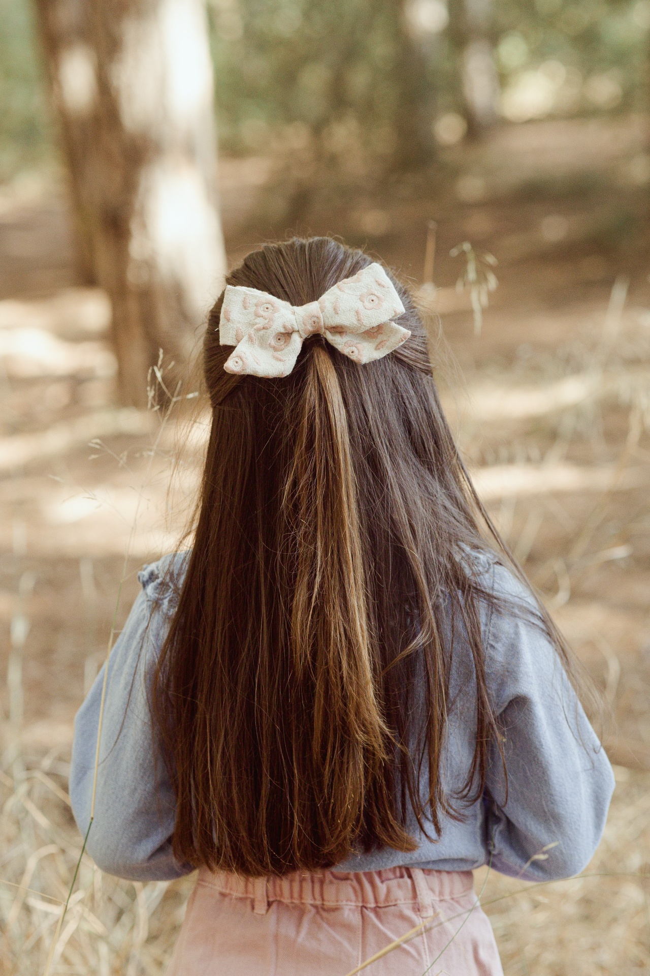 Laço de cabelo floral bege em cabelo castanho comprido de criança com roupa azul clara e rosa em ambiente natural