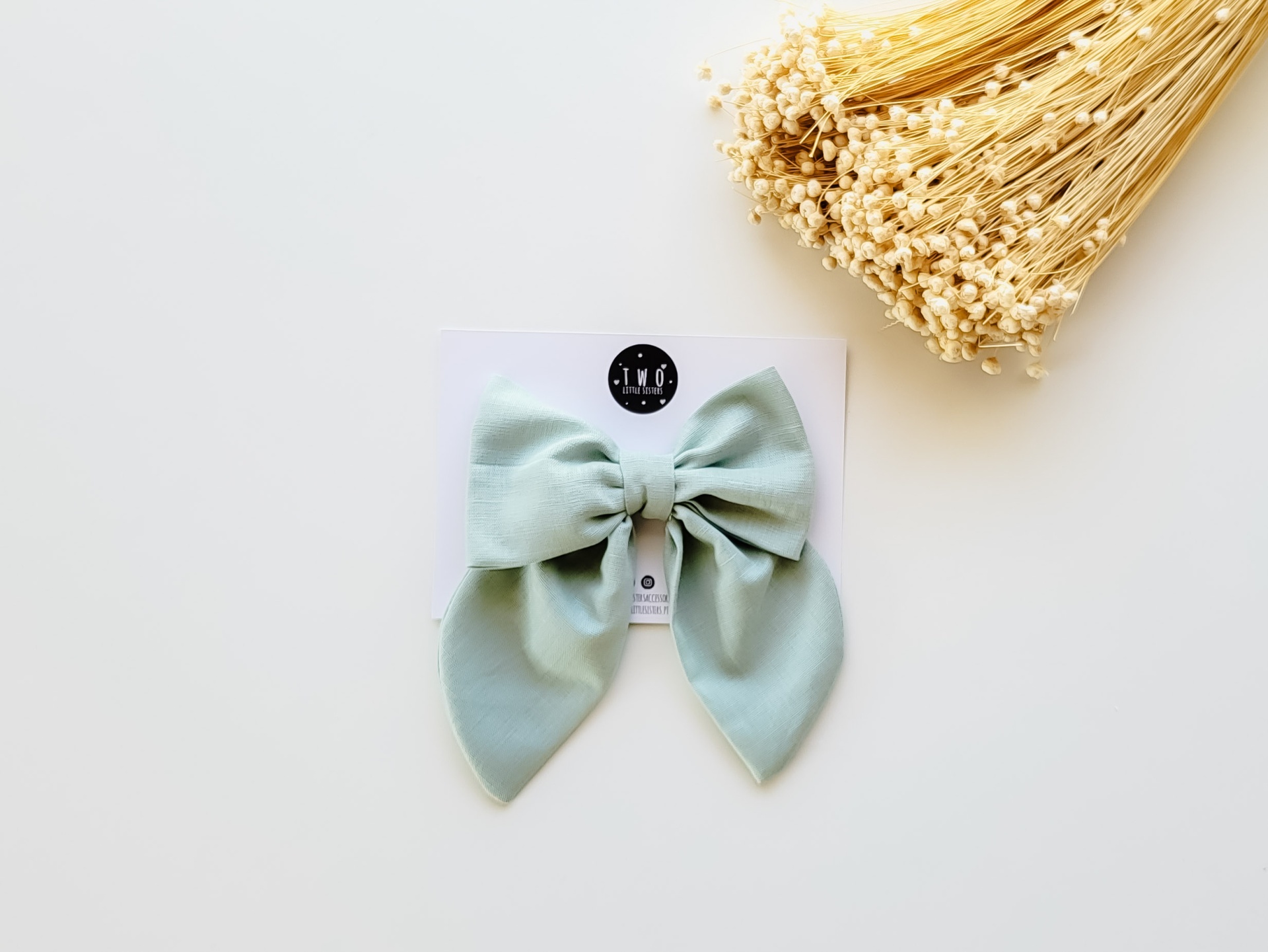 Laço tecido verde claro em cartão branco com logótipo T.N.T junto a flores secas amarelas