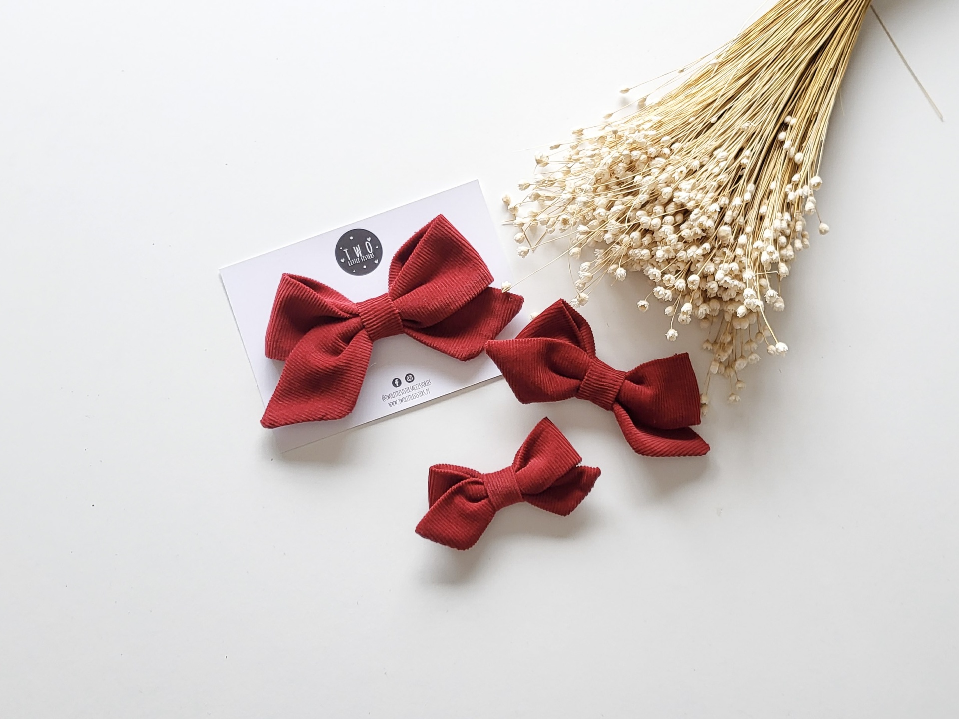 Três presilhas com laços vermelhos e ramo de flores secas sobre superfície branca