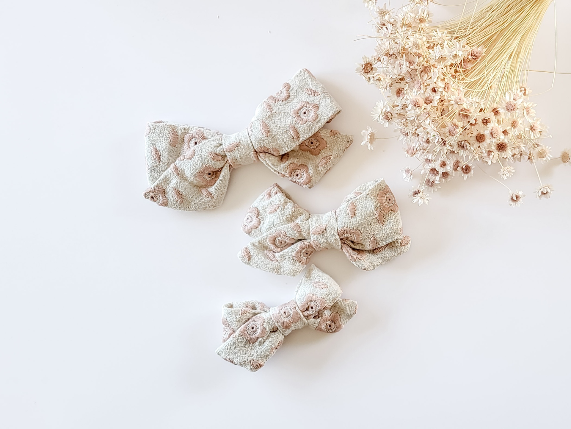 Três presilhas de cabelo com laços em tecido bege com padrão floral ao lado de flores secas sobre fundo branco