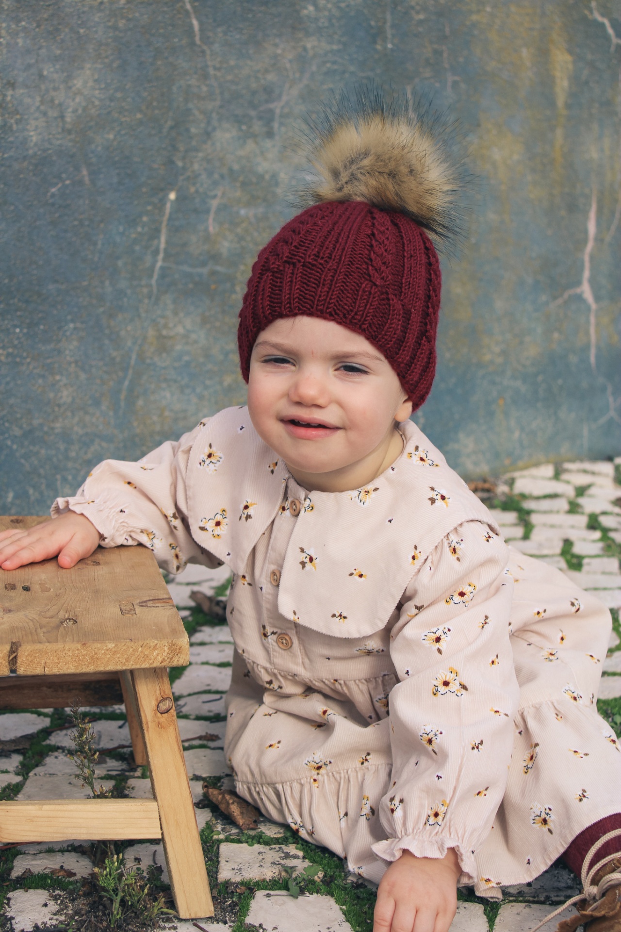 Criança com macacão floral bege e gorro vermelho com pompom sentada ao lado de banco de madeira