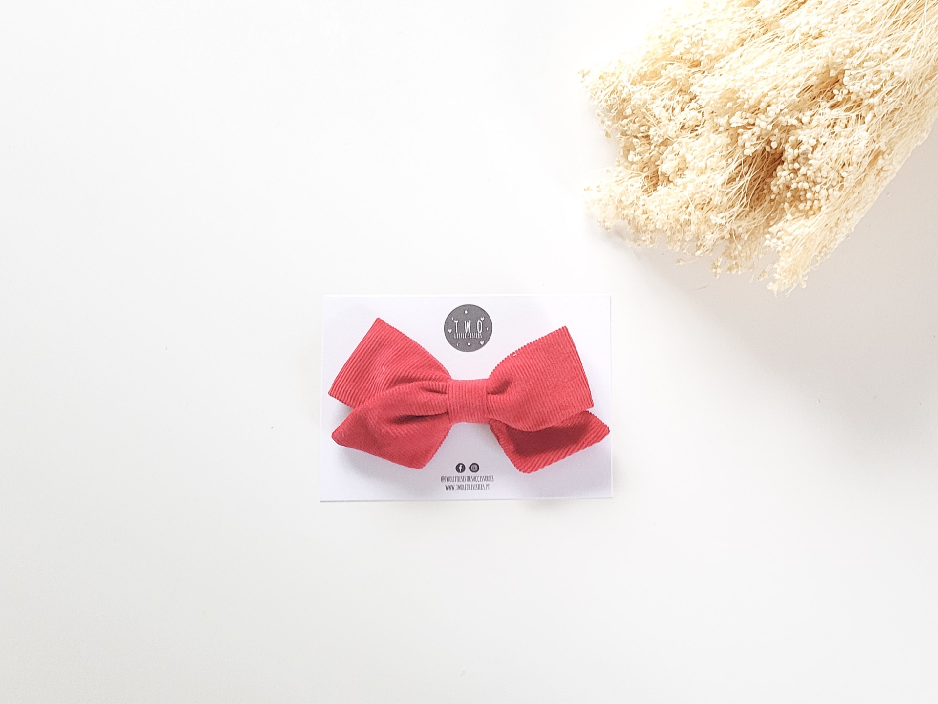 Laço vermelho em tecido fixado num cartão branco ao lado de flores secas