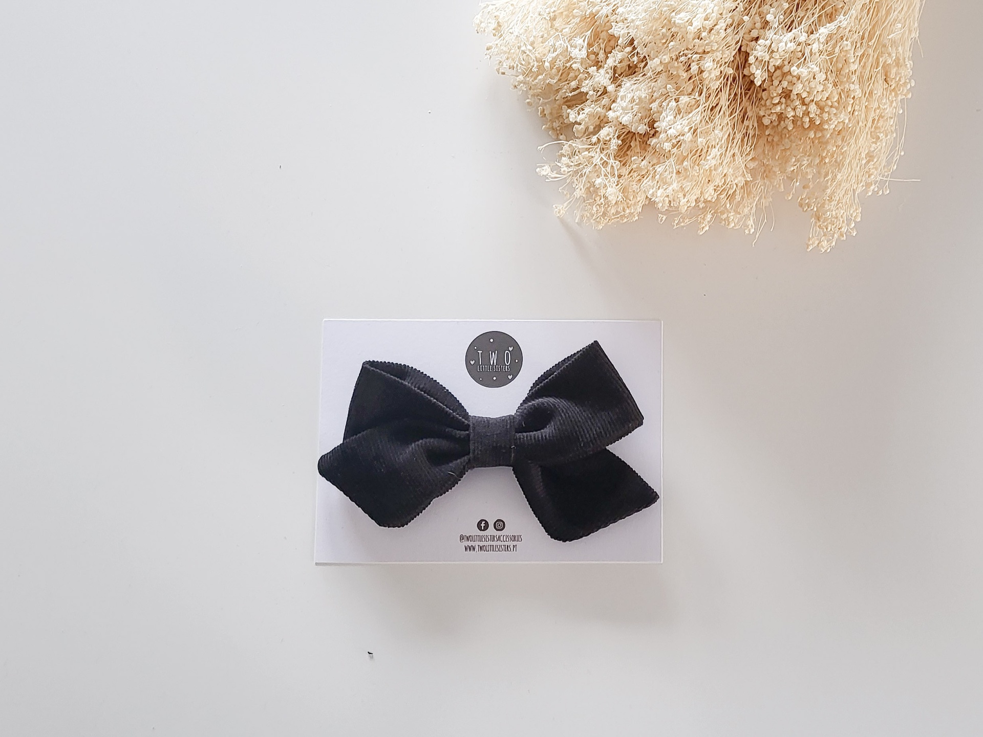 Laço preto em tecido canelado sobre cartão branco com logotipo e texto, flores secas no canto