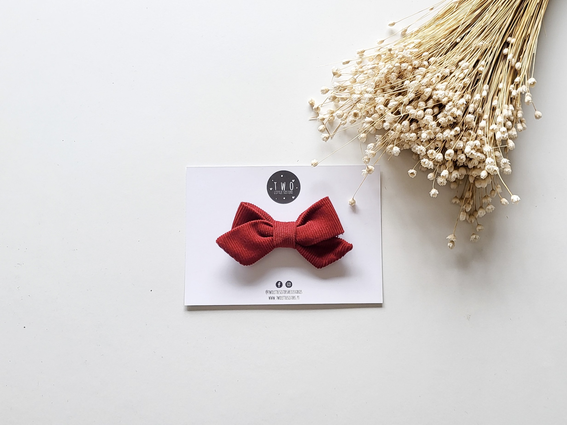 Laço vermelho de tecido canelado sobre cartão branco com texto, ao lado de flores secas brancas.