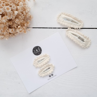 Quatro molas de cabelo brancas de tecido frisado, duas num cartão e duas soltas numa mesa branca com flores secas ao lado.