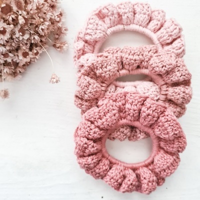 Três scrunchies de crochê rosas em superfície branca com flores secas ao lado
