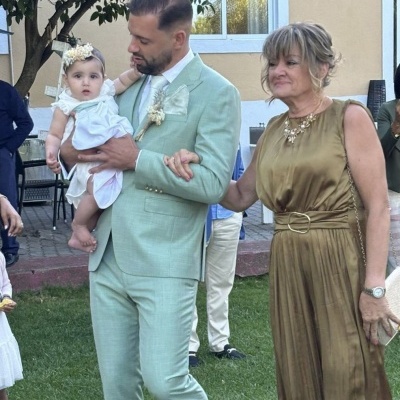 Homem de fato verde claro segura criança com vestido branco ao lado de mulher de vestido dourado em jardim