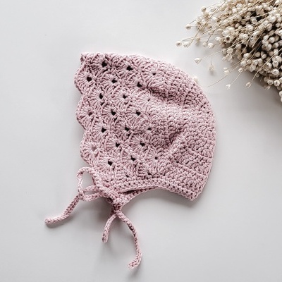 Boné infantil rosa claro de croché com padrão rendado e fitas ao lado de flores secas