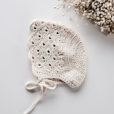 Gorro de malha cru com padrão vazado e fitas, ao lado de flores secas brancas.