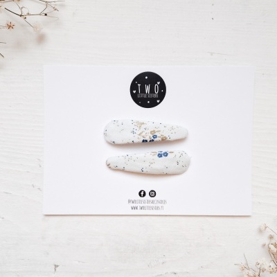 Presilhas de cabelo brancas com manchas azuis e beges sobre cartão branco com logótipo preto, em fundo branco com flores secas.