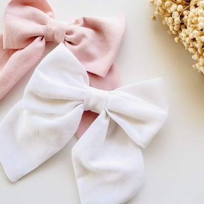 Dois laços de tecido, branco e rosa, sobre superfície branca com flores secas ao lado