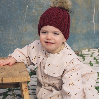 Criança com macacão floral bege e gorro vermelho com pompom sentada ao lado de banco de madeira