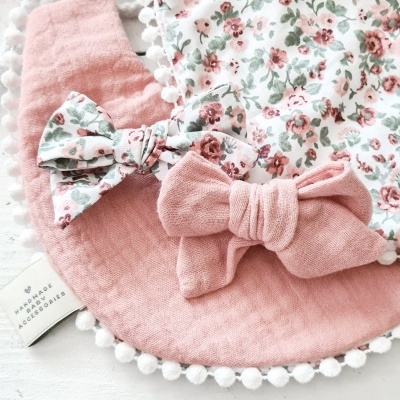 Babetes de bebé em tecido com padrão floral e pompons e laço rosa sobre mesa branca