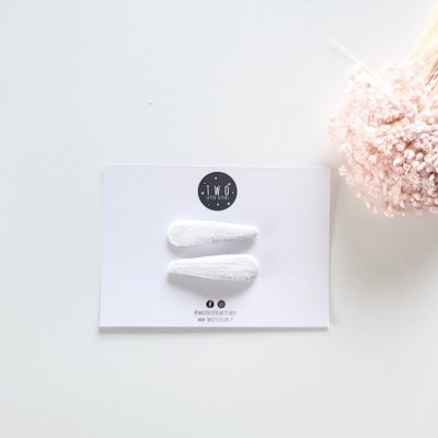 Dois ganchos de cabelo brancos em cartolina branca com logotipo da marca e flores cor-de-rosa ao lado