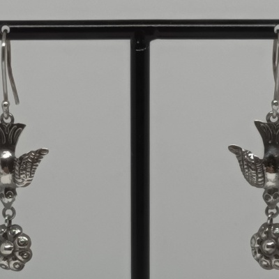 Brincos prateados com desenho de pássaro e detalhe circular, pendurados num suporte preto.