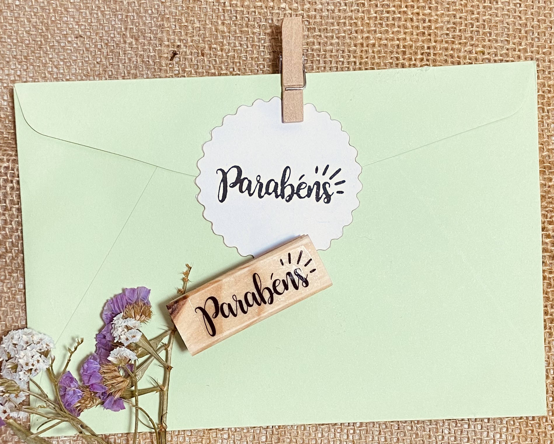 Envelope verde pálido com autocolante 'Parabéns', pregadores de madeira e flores secas roxas e brancas