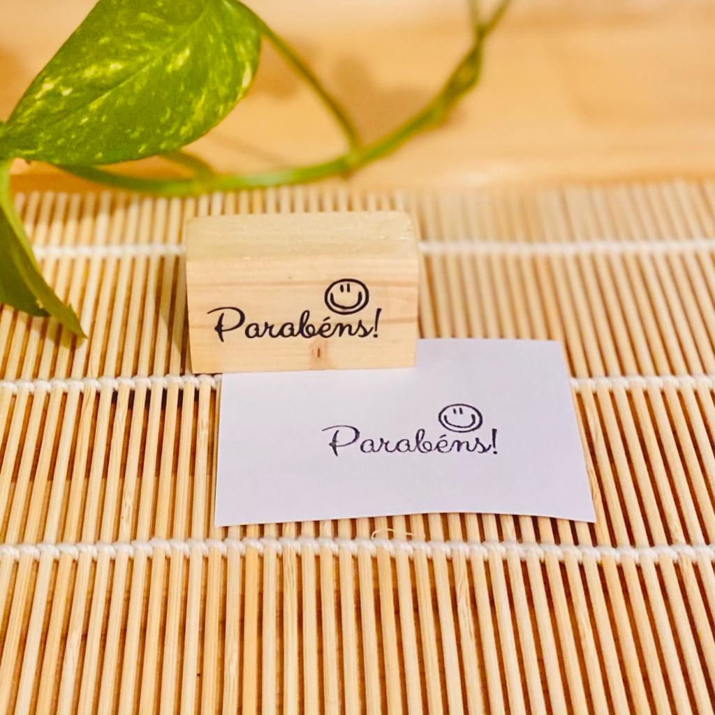 Carimbo de madeira com texto 'Parabéns!' e um sorriso