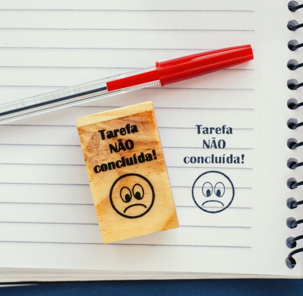 Selo de borracha com texto 'Tarefa NÃO concluída!' e cara triste, caneta vermelha e papel de linhas