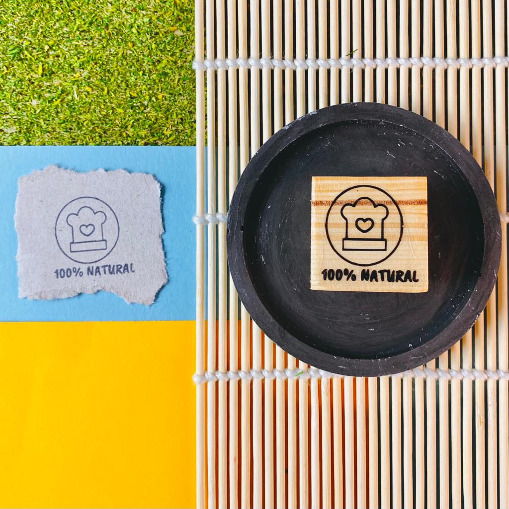 Bloco de madeira com carimbo 100% NATURAL em esteira de bambu ao lado de papel com mesma imagem e fundo colorido.