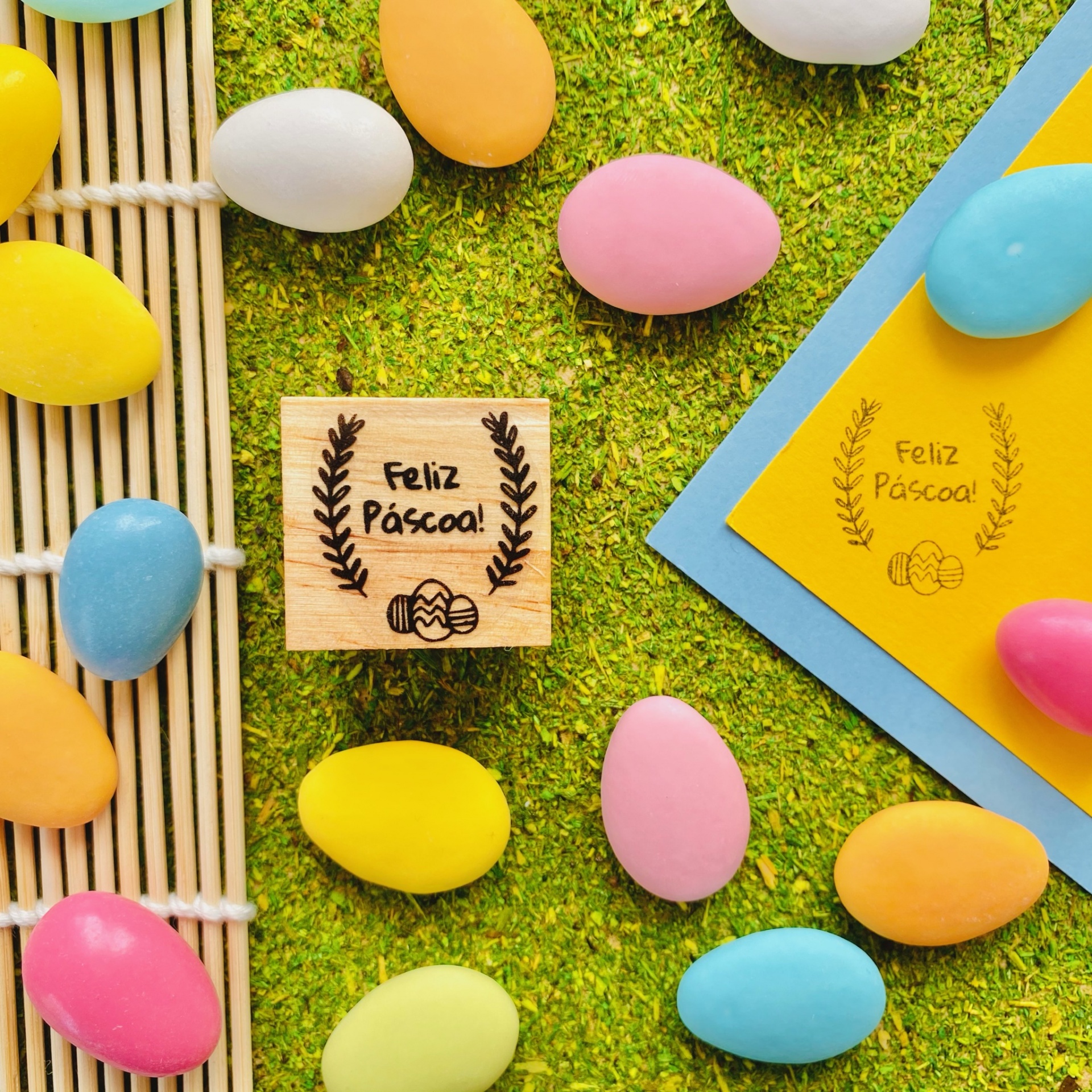 Carimbo de madeira com frase Feliz Páscoa rodeado por ovos coloridos sobre superfície verde