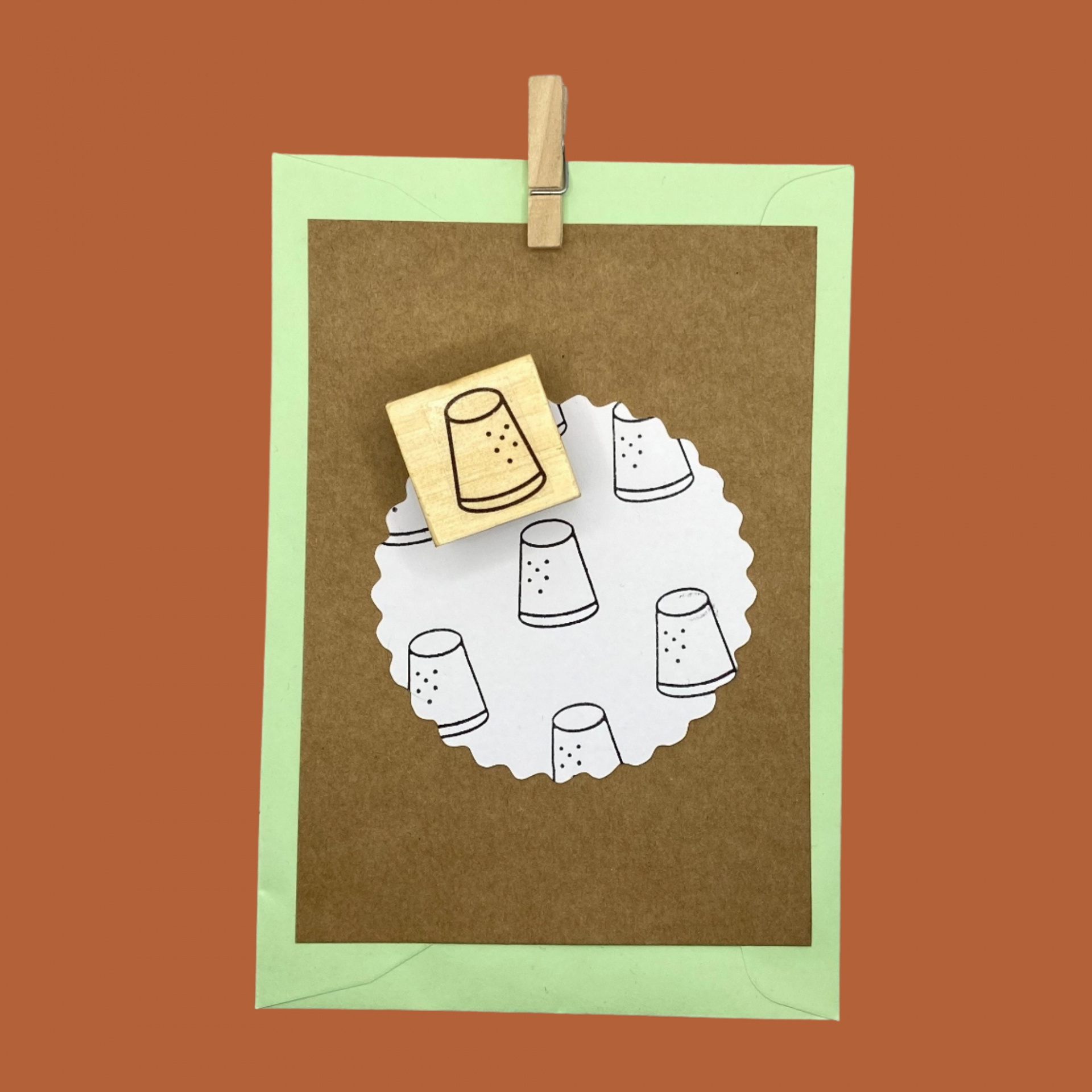 Carimbo de madeira com desenho de dedal preto sobre papel branco em círculo e cartão castanho e verde