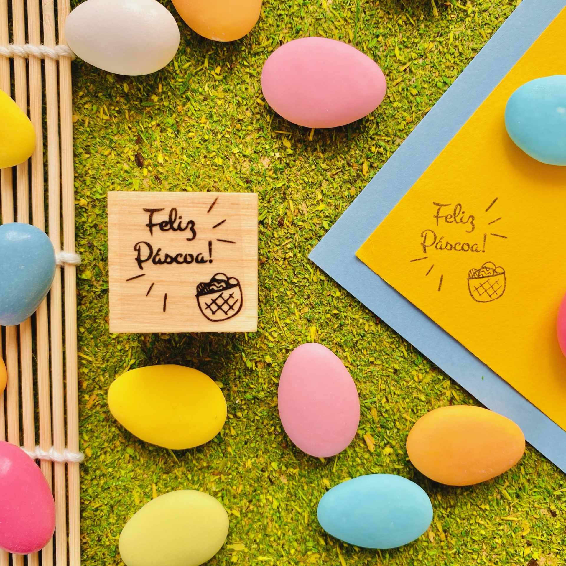Carimbos de madeira com texto Feliz Páscoa junto a ovos de Páscoa coloridos sobre grama verde.