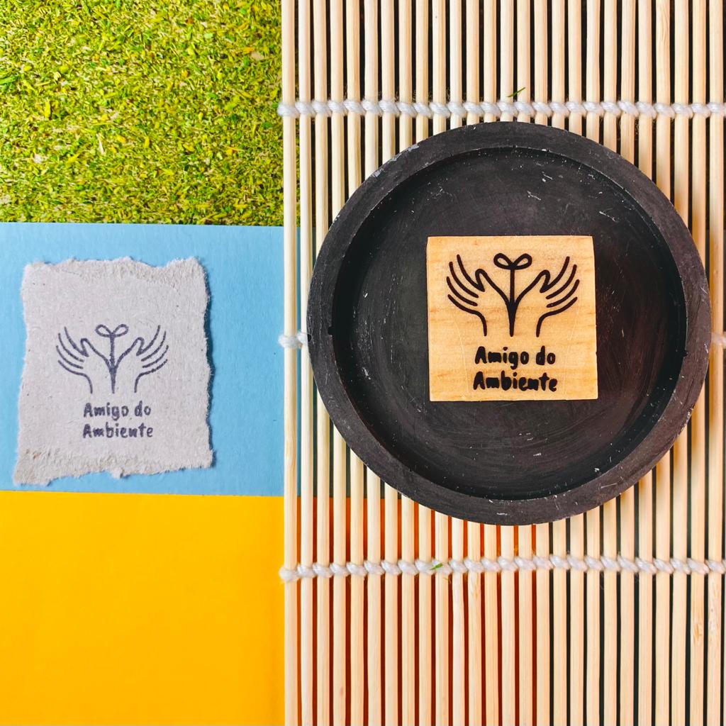 Carimbo de madeira com desenho de planta e mãos e a frase Amigo do Ambiente em tigela preta sobre esteira de bambu