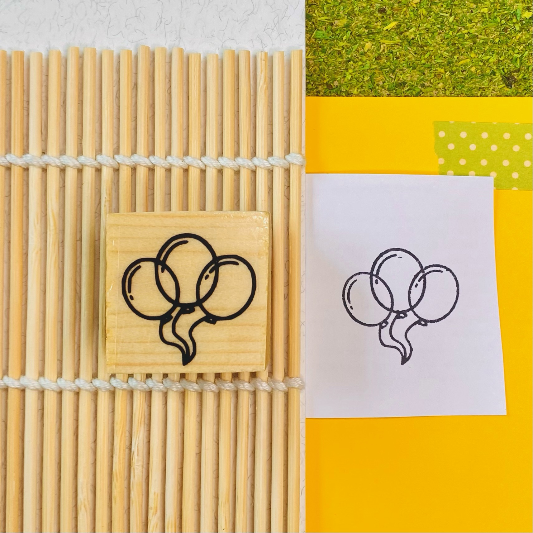 Carimbo de balões em madeira sobre esteira de bambu e papel com a impressão do carimbo numa superfície amarela