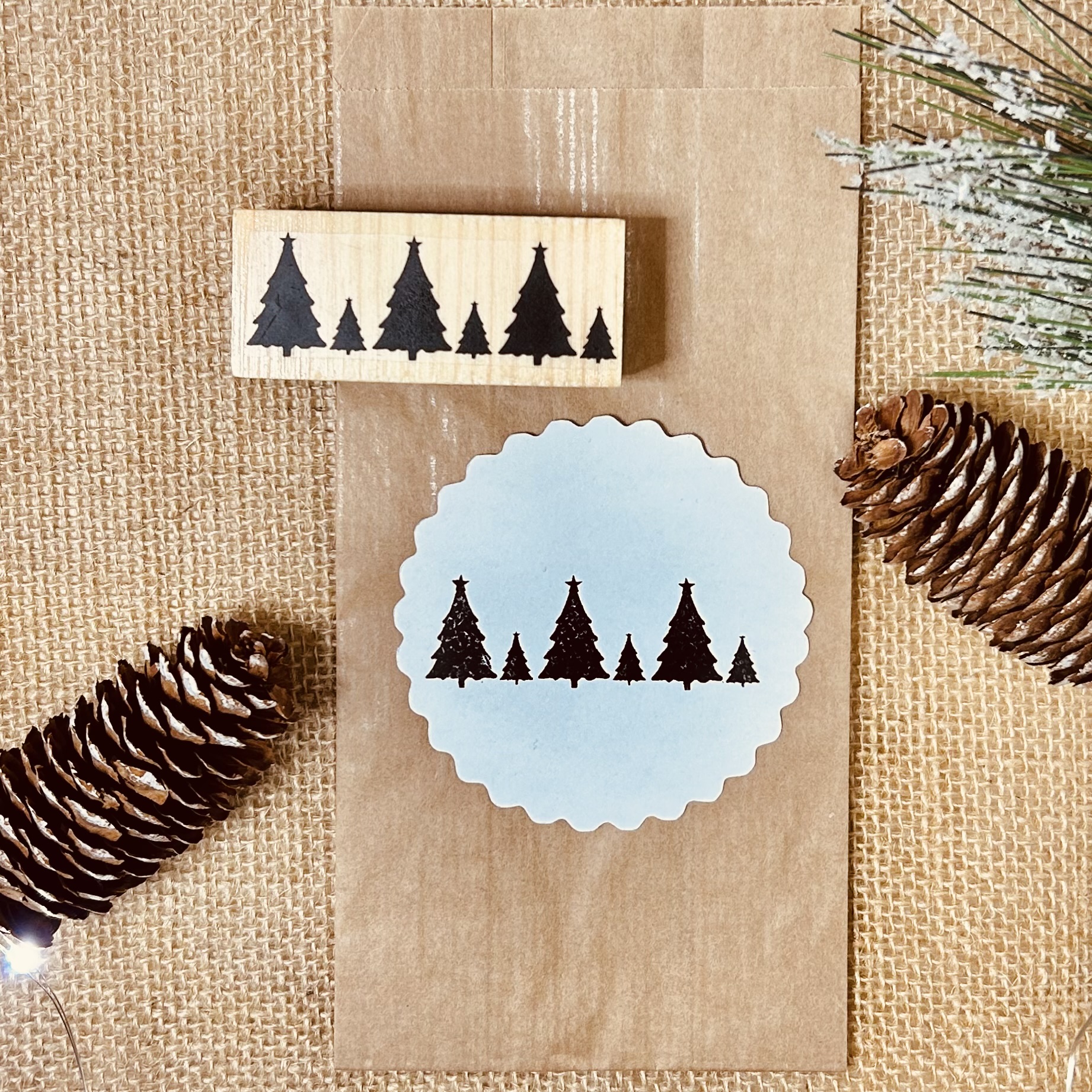 Carimbo de madeira com figuras de árvores de Natal e círculo de papel estampado sobre tecido castanho com pinhas e ramos secos