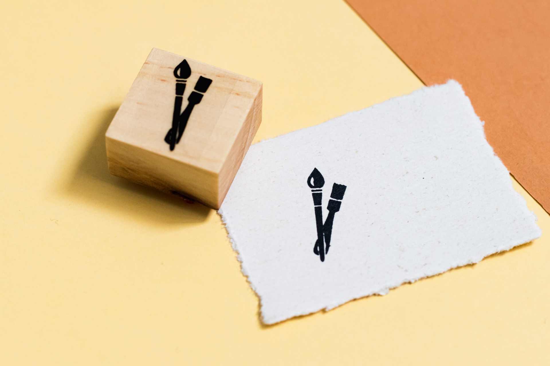 Carimbo de madeira quadrado com desenho de pincéis cruzados e papel branco com o mesmo desenho sobre fundo amarelo