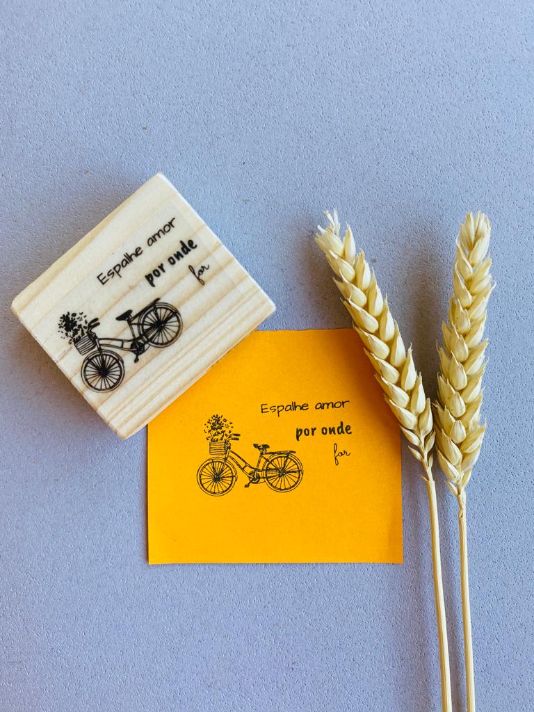 Carimbo de madeira com desenho de bicicleta e texto, impressão em papel laranja e três espigas de trigo sobre fundo cinzento