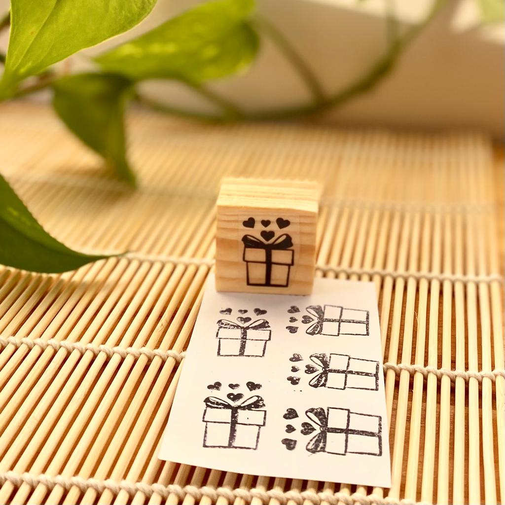 Carimbo de madeira com desenho de presente com corações e papel branco com estampas do carimbo sobre superfície de bambu.