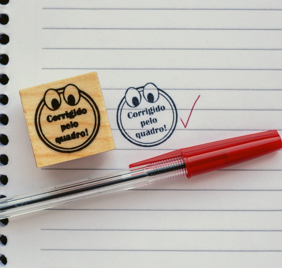 Carimbo de madeira com texto 'Corrigido pelo quadro!' e caneta vermelha sobre caderno pautado