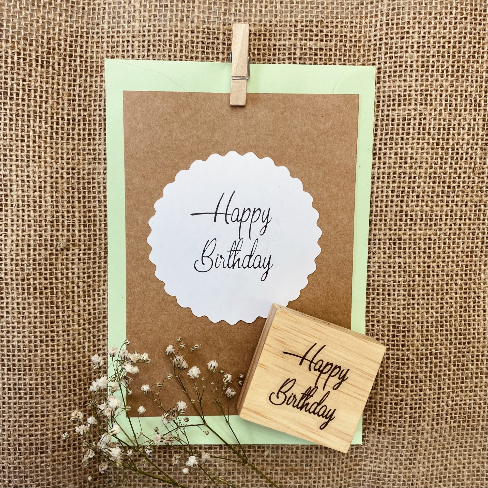 Cartão de aniversário com texto Happy Birthday, selo de borracha e flores secas sobre fundo de tecido rústico