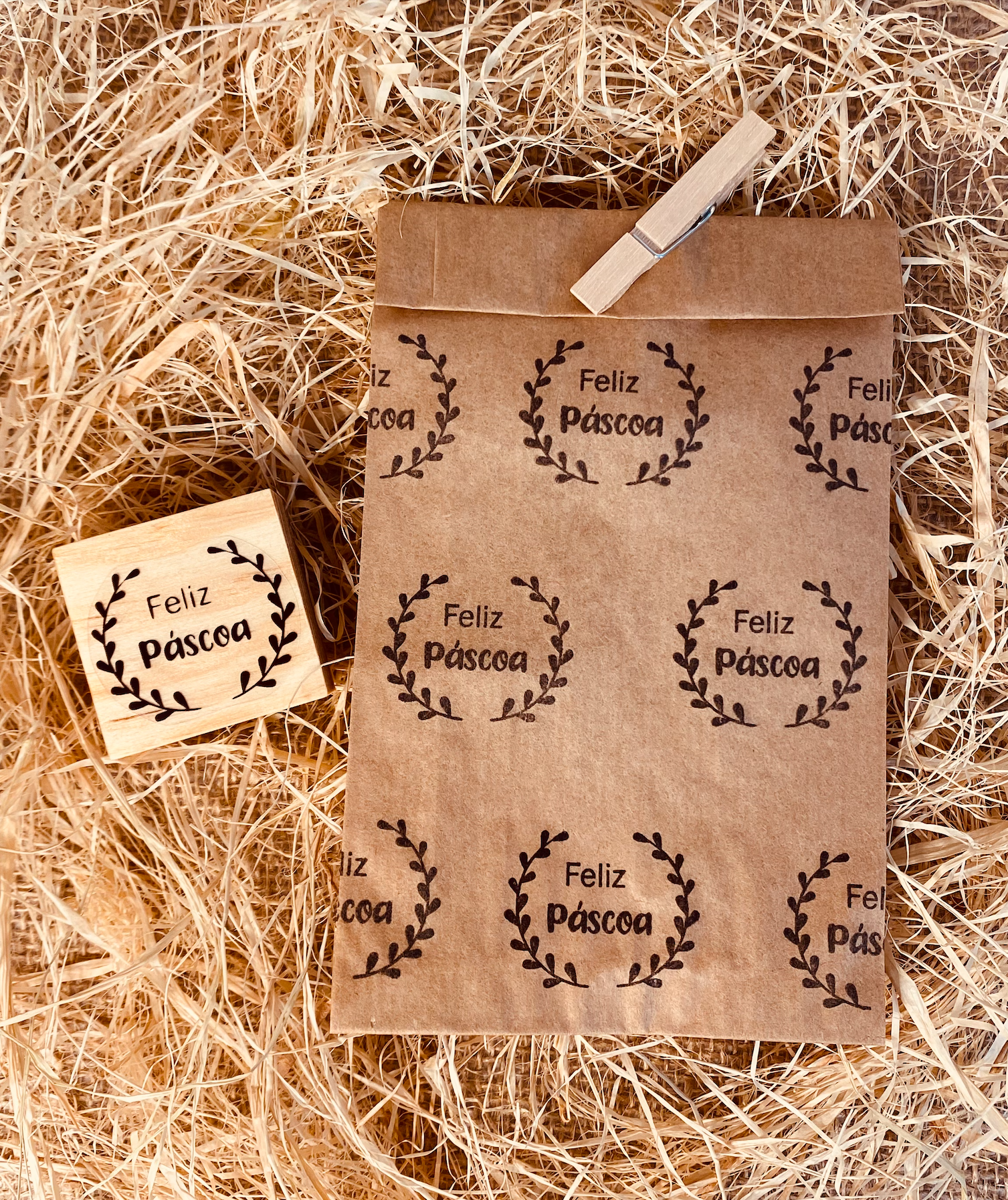 Selo de borracha com carimbo e saco de papel com impressão 'Feliz Páscoa' em tinta preta sobre fundo de palha.