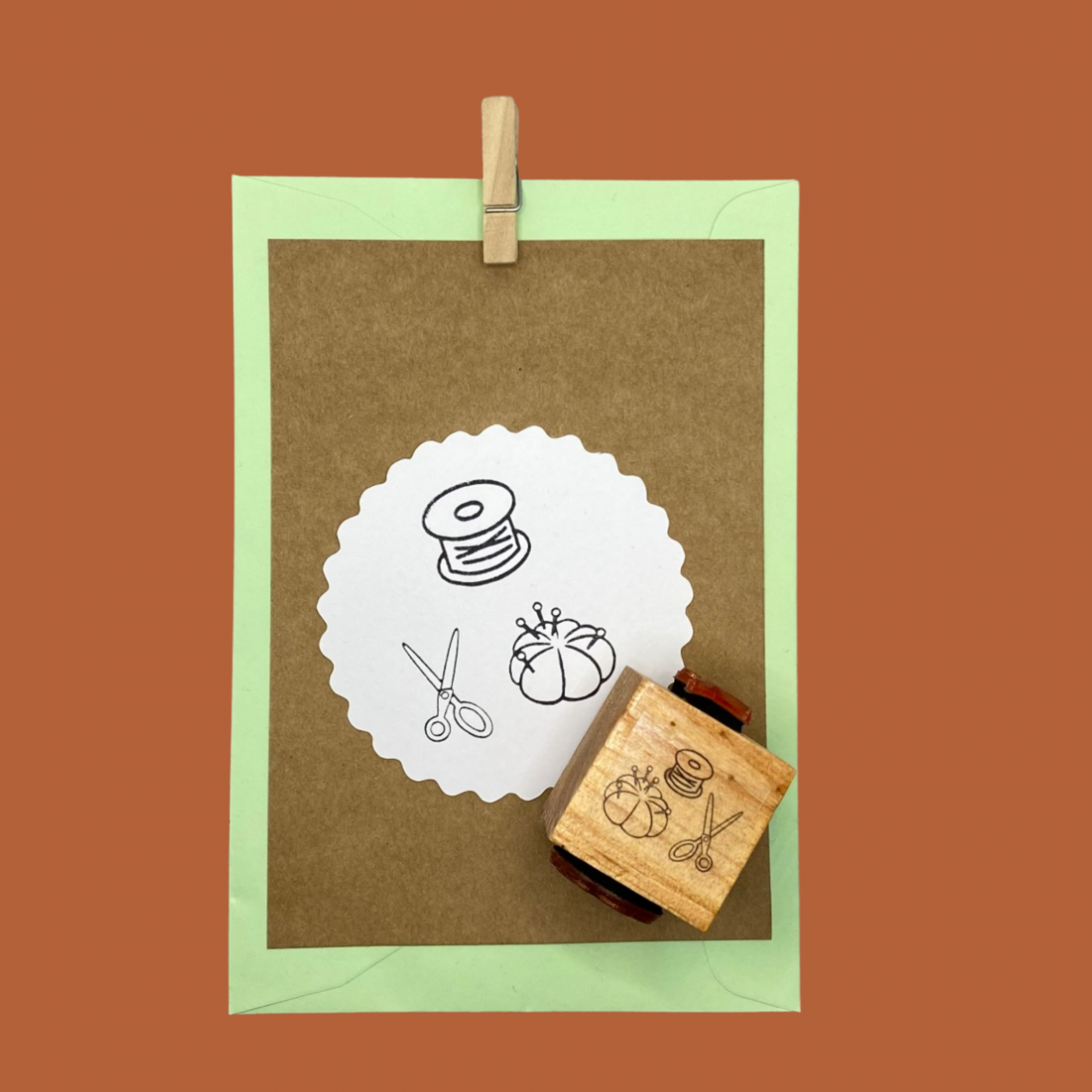 Carimbo de madeira com desenho de linha de tesoura e almofada de alfinetes num papel castanho em envelope verde