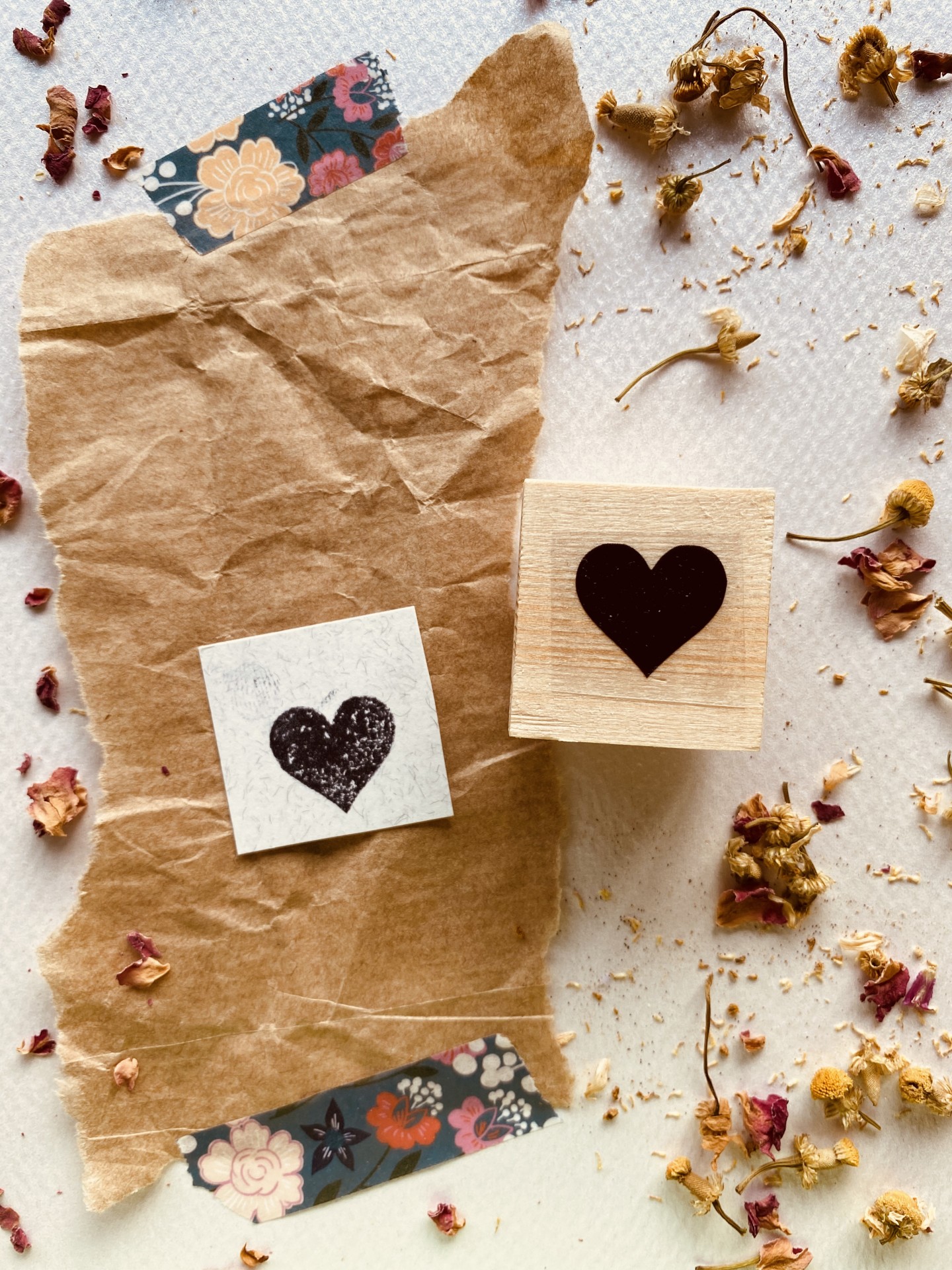 carimbo de madeira com coração preto, papel castanho, fita adesiva floral, flores secas sobre fundo branco