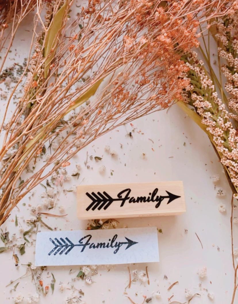 Carimbo de borracha com a palavra 'Family' e uma seta preta, com flores secas ao redor