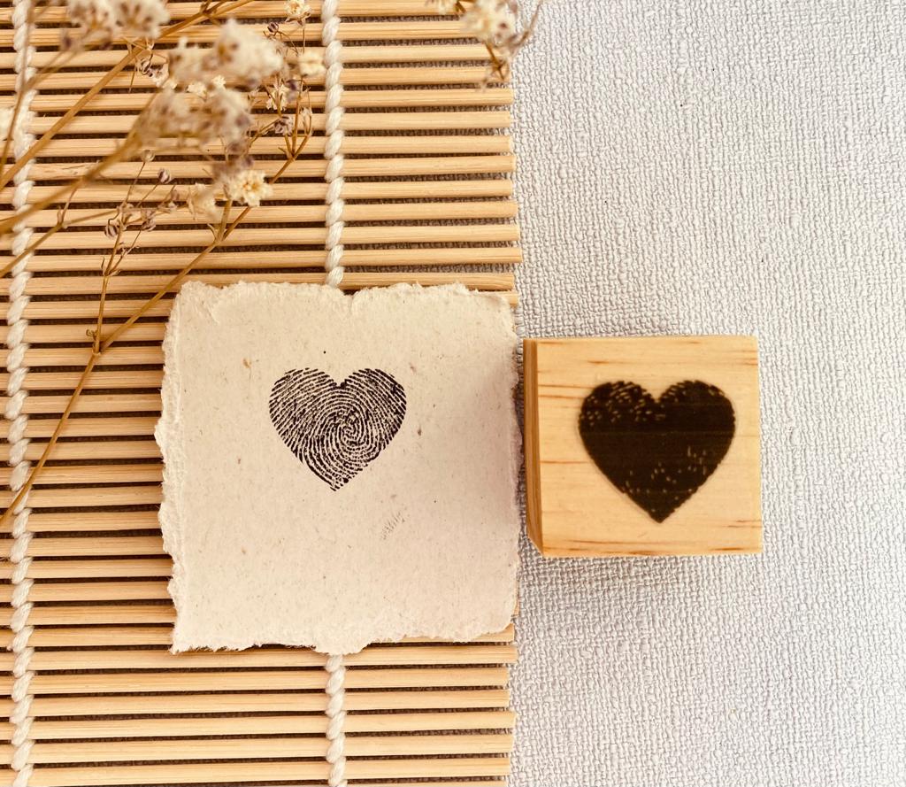 Carimbo de coração em madeira e papel artesanal com estampa de coração preta sobre tapete de bambu e flores secas