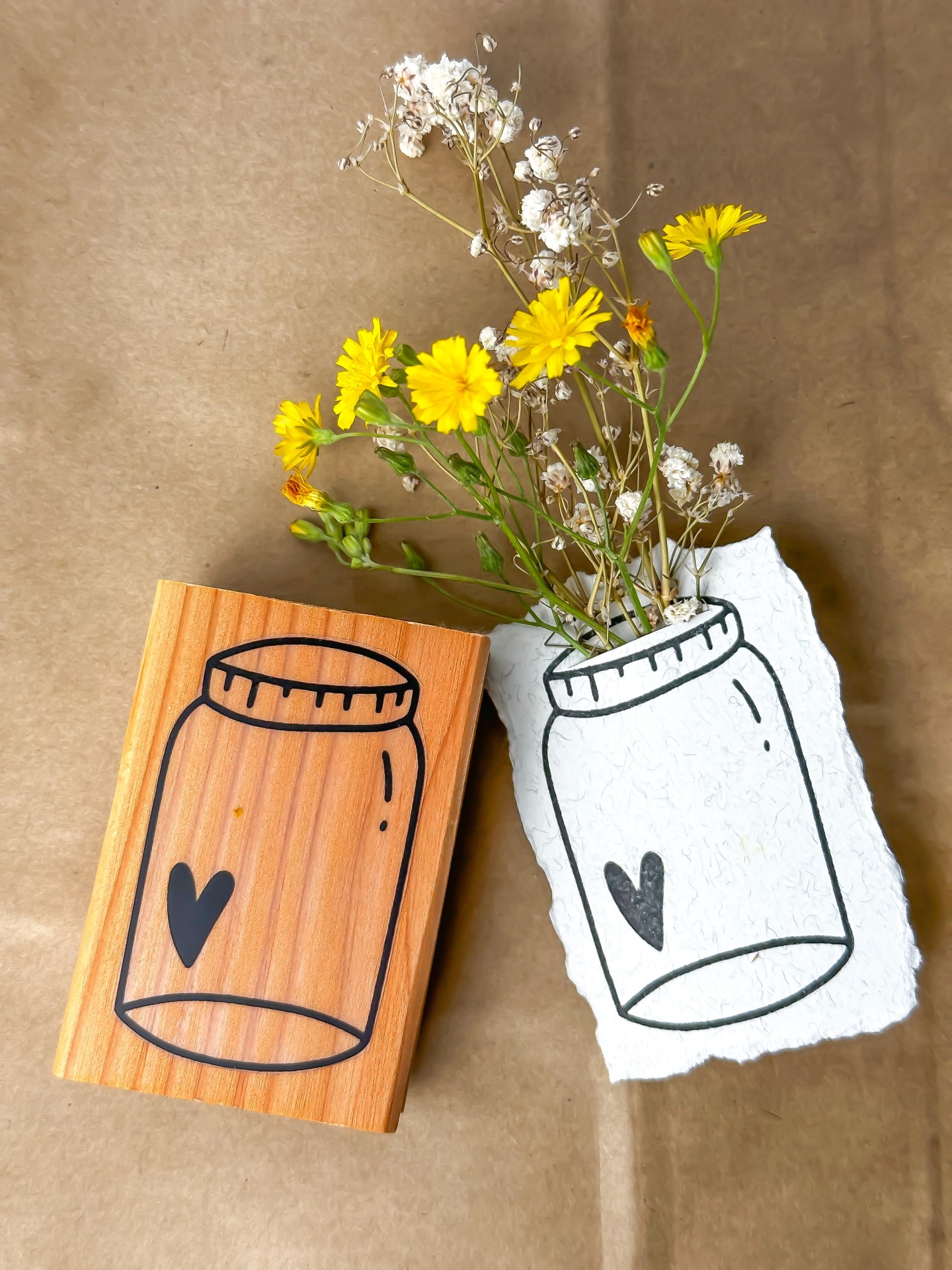 Carimbo de madeira com desenho de frasco e coração com impressão em papel e flores amarelas.