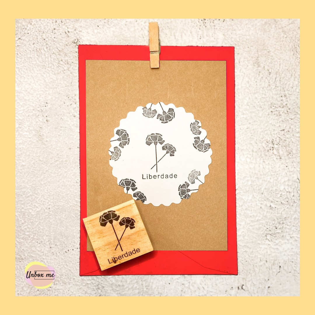 Cartão kraft com envelope vermelho, etiqueta branca com flores e texto 'Liberdade' e carimbo de madeira