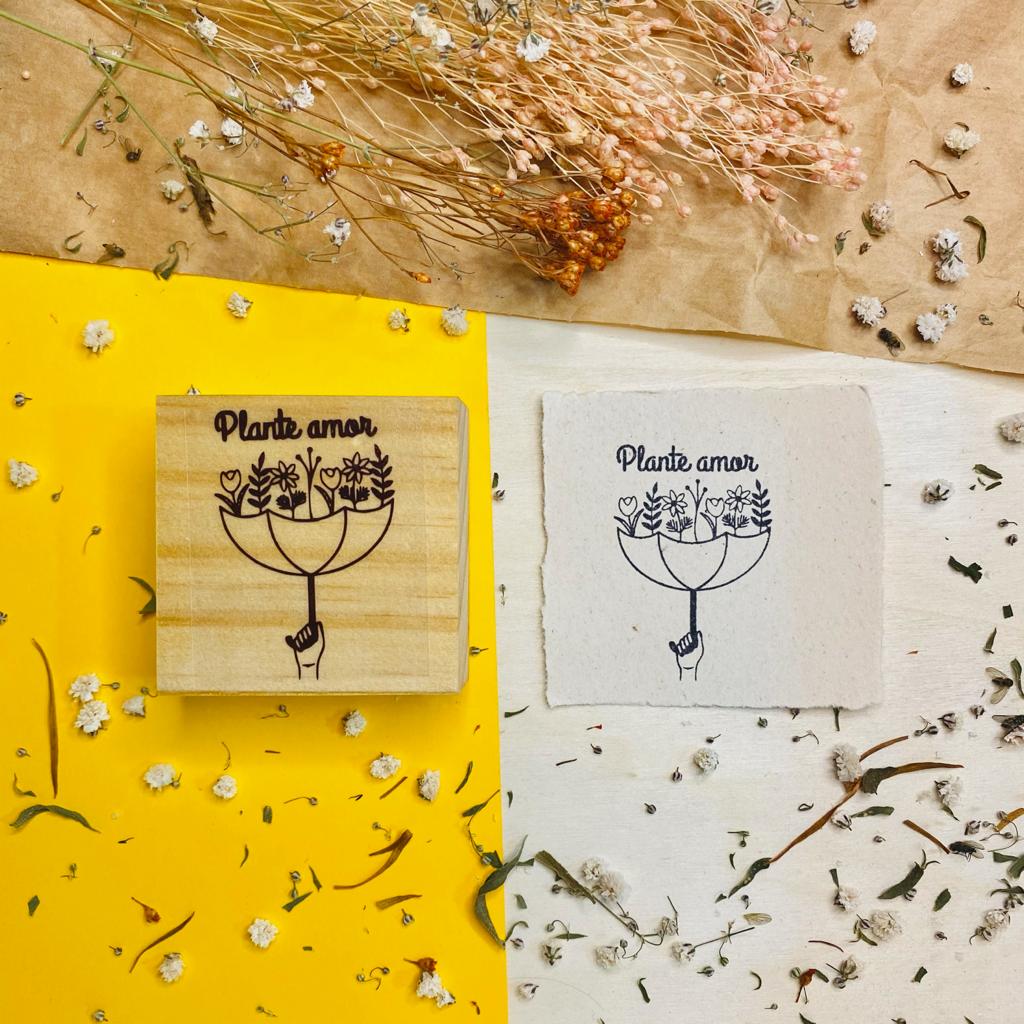 Selo de madeira com estampa 'Plante amor' ao lado de papel branco com a mesma estampa, em fundos amarelo e branco com flores secas.