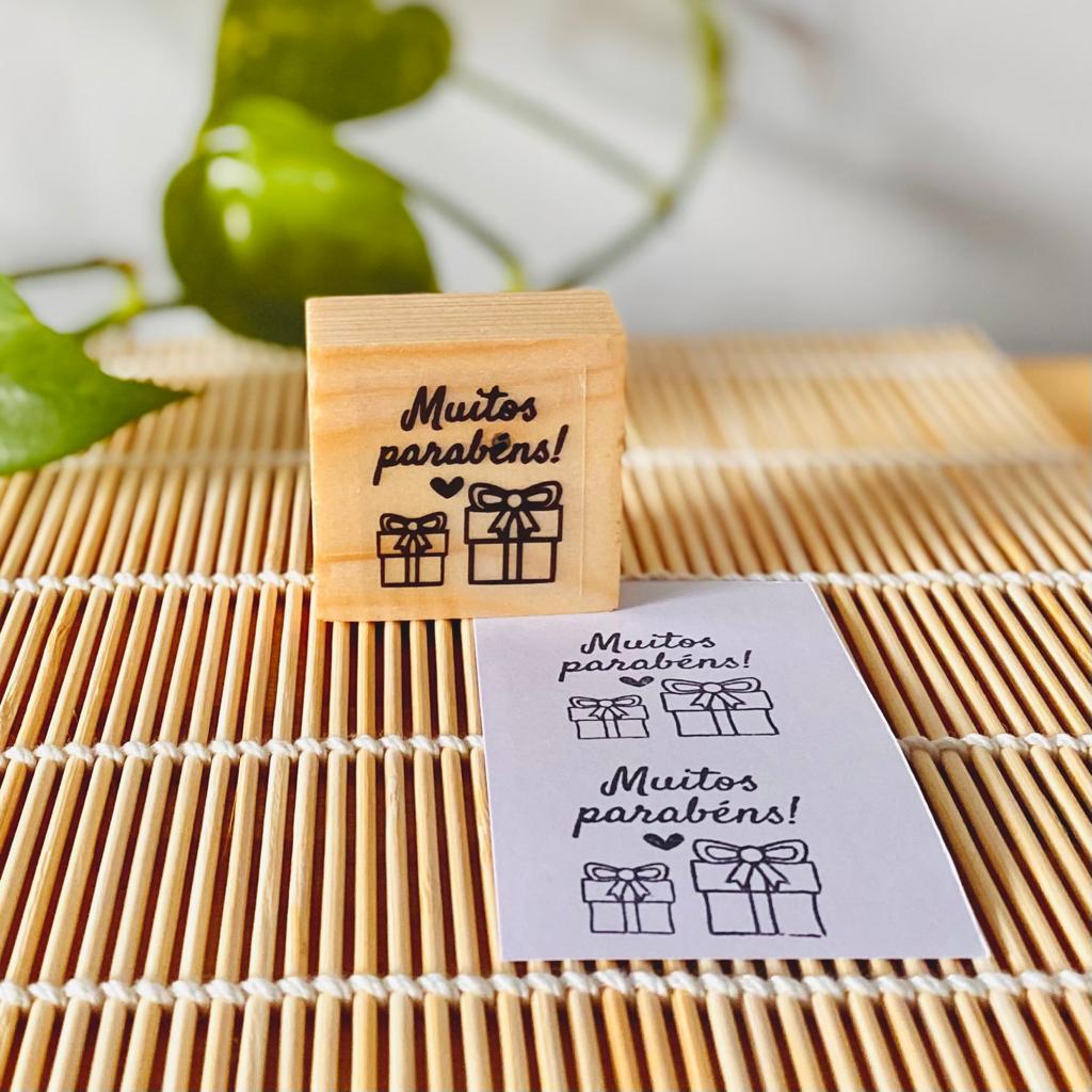 Carimbo de madeira com texto e ícones de caixas de presente sobre tapete de bambu com um papel carimbado e planta ao fundo