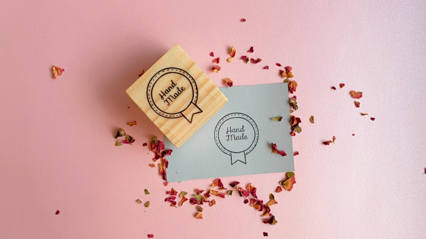Selo de madeira com texto 'Hand Made' sobre fundo rosa com papel cinzento e pétalas secas