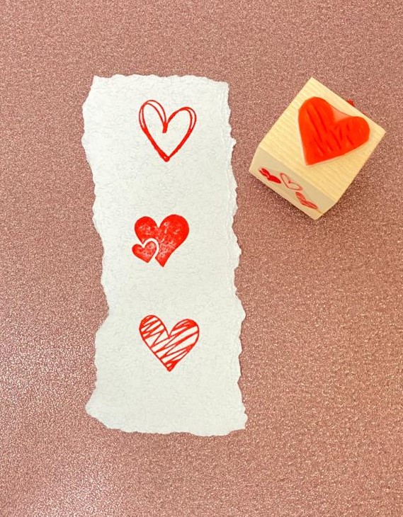 Carimbo de madeira com coração vermelho e impressões em papel branco sobre fundo rosa