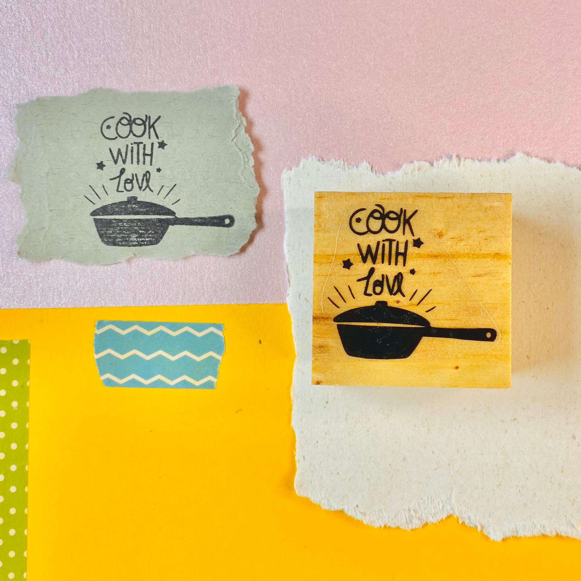 Selo de borracha amarelo com desenho de frigideira e texto 'COOK WITH LOVE' sobre fundo colorido