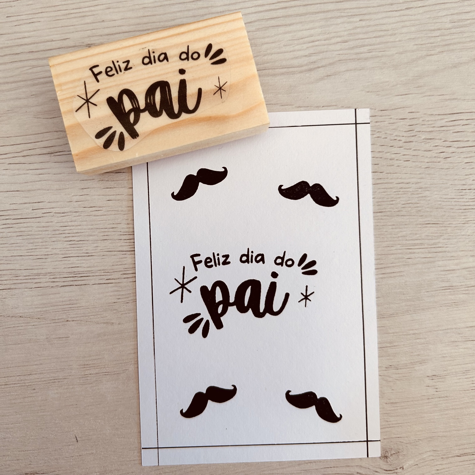 Carimbo de madeira com texto 'Feliz dia do pai' e cartão branco com mesma frase e desenhos de bigodes