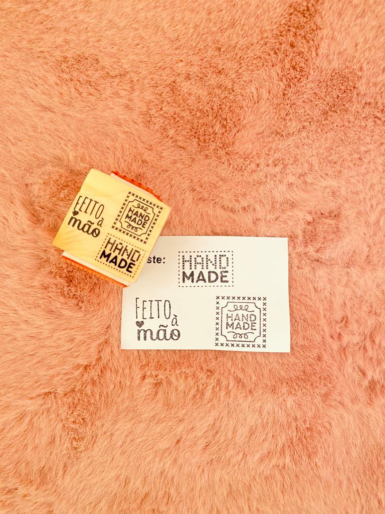 Carimbo de madeira e borracha com textos 'FEITO à mão' e 'HAND MADE' sobre superfície felpuda rosa