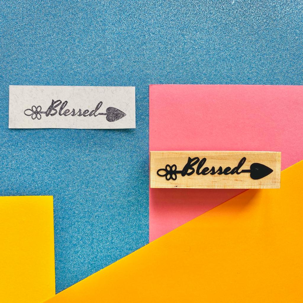 Carimbo de madeira com texto Blessed e sua impressão numa etiqueta branca sobre fundo colorido