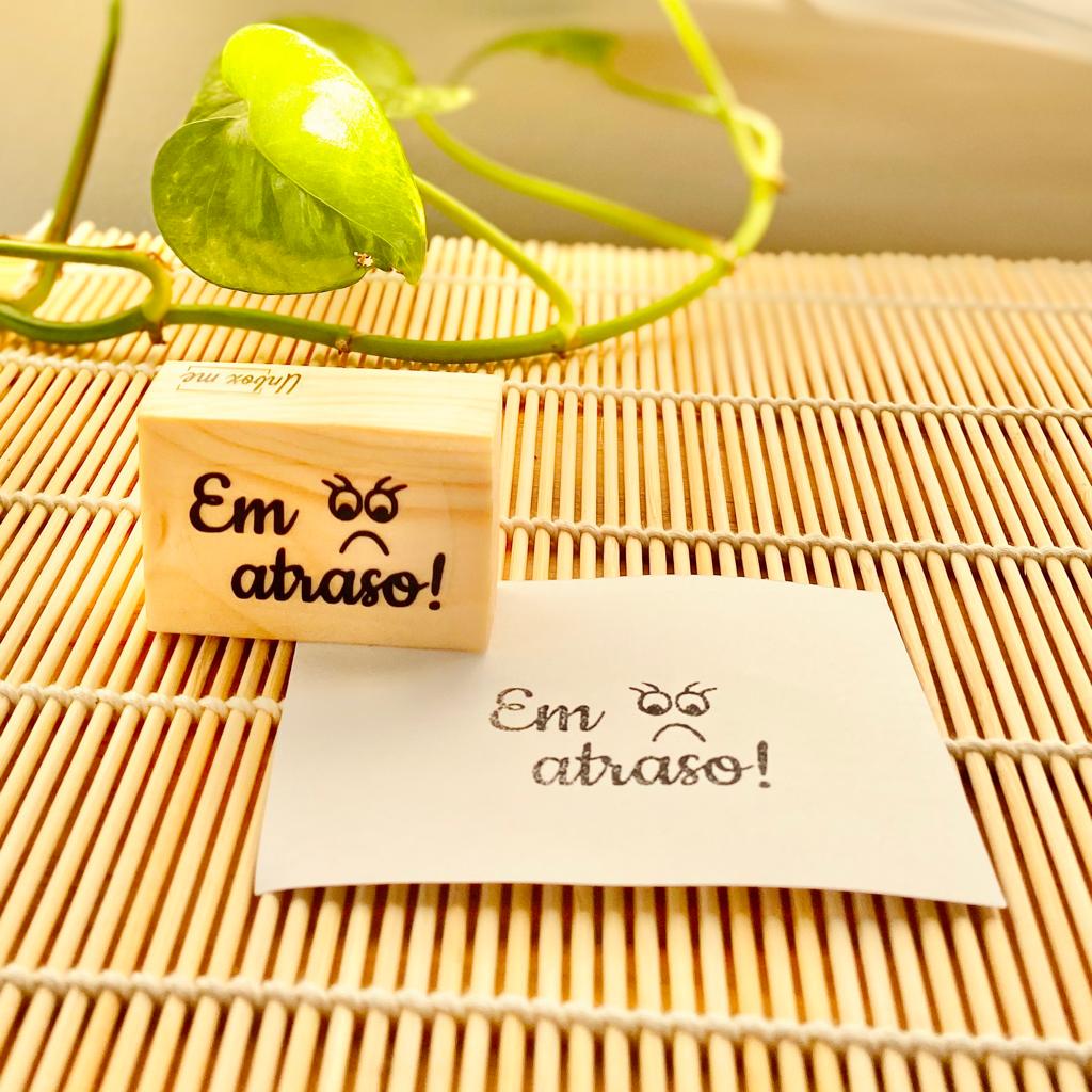 Carimbo de madeira com a frase 'Em atraso!' e olhos irritados, junto com impressão em papel sobre tapete de bambu